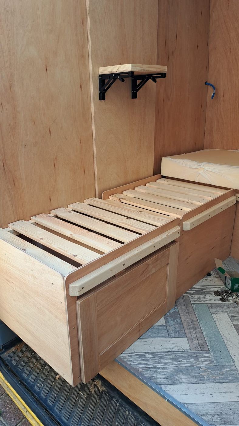 Sofa bed base carpentry with slatted top and fold-down shelf bracket inside a campervan
